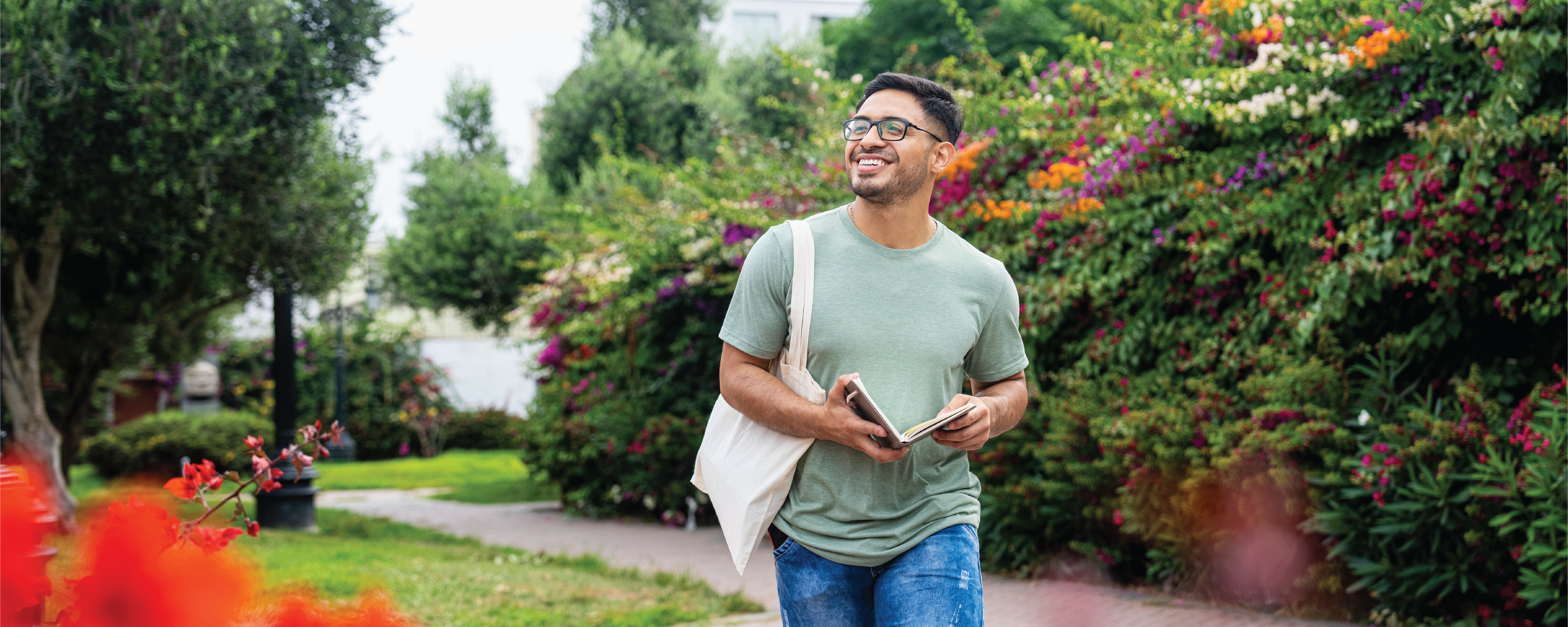 A smiling person wearing glasses, a light green T-shirt, and blue jeans walks along a garden path while holding a notebook. They carry a white tote bag over one shoulder and are surrounded by lush greenery and colorful flowering shrubs. Bright red flowers frame the foreground, and a pigeon stands on the path in the lower right corner.