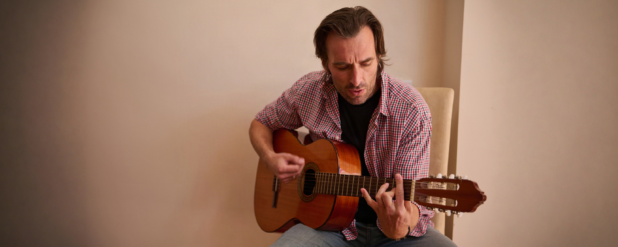 Man seated indoors playing an acoustic guitar, wearing a red plaid shirt over a black T-shirt.