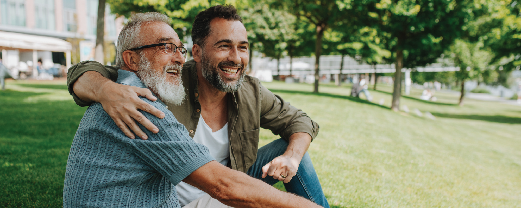 An older man with a white beard and glasses and a younger man sit together on the grass in a park, smiling and embracing as they enjoy time outdoors on a sunny day.