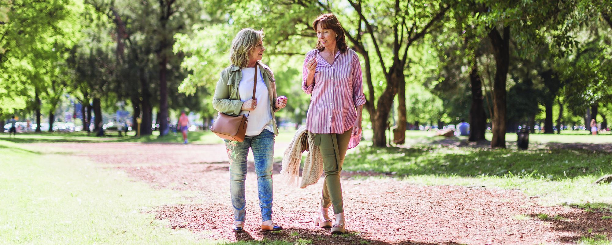 Two women walk side by side along a sunlit park path, smiling and talking while carrying light jackets. Tall trees cast dappled shade over the trail, creating a warm, relaxed outdoor setting.