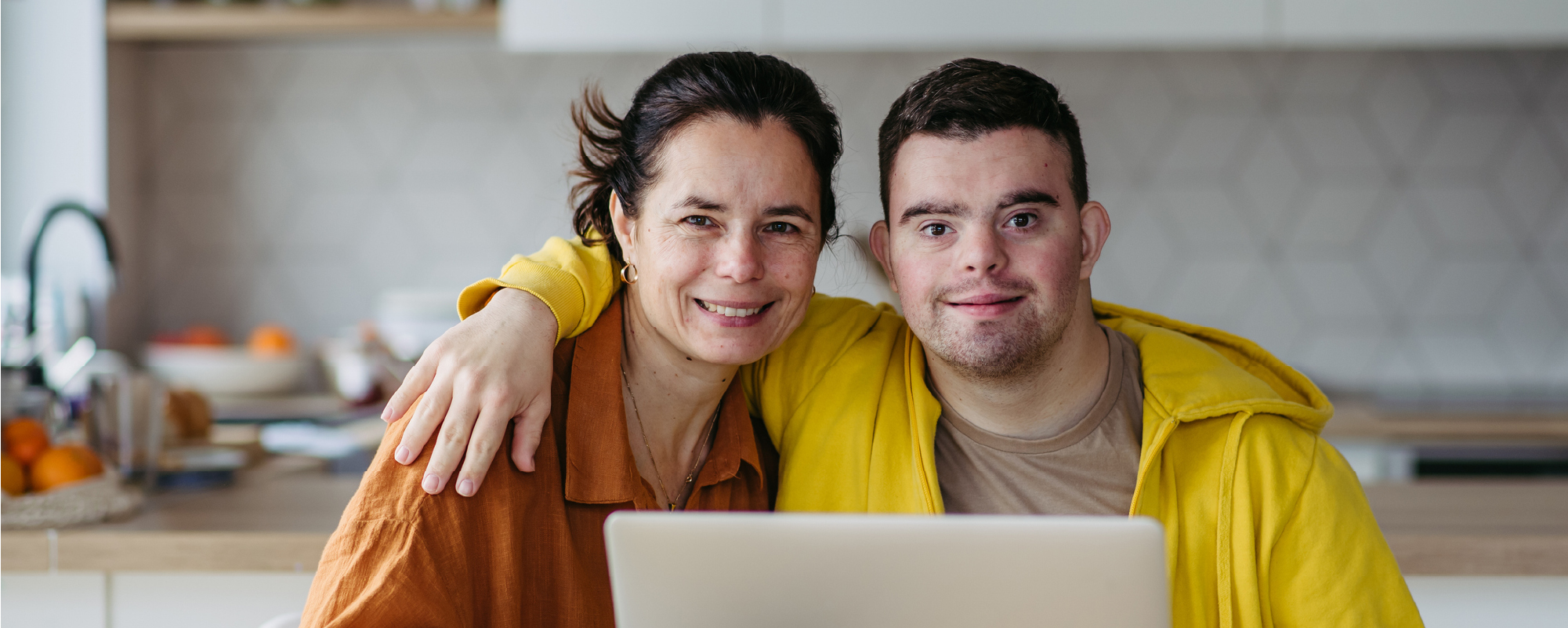 Woman and young man sitting close together at a kitchen table, smiling at the camera behind an open laptop.