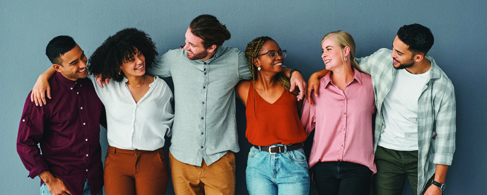 Six smiling adults of diverse backgrounds stand side by side with their arms around each other's shoulders, sharing a friendly moment against a plain blue wall. They wear casual clothing and look at one another, conveying connection, support, and inclusivity.